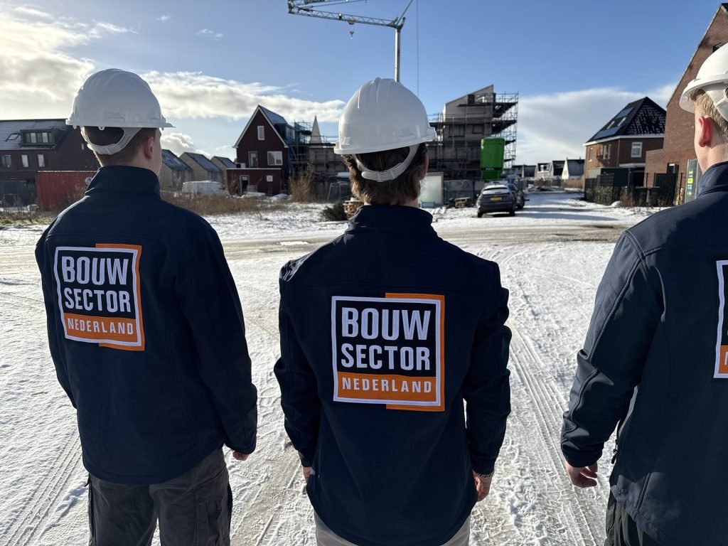Drie vakmensen van Bouwsector Nederland met witte helm op een besneeuwde straat met woningen in aanbouw en een bouwkraan